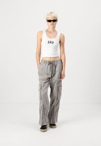 Bijela skraćena majica bez rukava s "GAP" logotipom u tamnoplavoj boji, uparena s sivim cargo hlačama s patentnim zatvaračima i bočnim džepovima, upotpunjena crnim cipelama.
