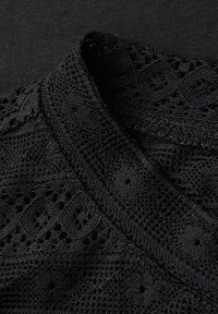 Tessuto nero con colletto in pizzo intricati; presenta ritagli geometrici e motivi testurizzati; materiale di base liscio sotto il dettaglio in pizzo.