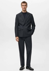 Chaqueta de traje cruzada color gris oscuro con pantalones a juego. Tejido liso, diseño estructurado y elegantes zapatos negros que completan el conjunto.