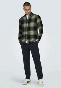 Chemise en flanelle à carreaux noire et verte à manches longues, avec un design à boutons à l'avant, associée à un pantalon bleu marine et des baskets blanches.