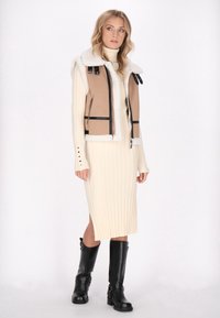 Beige schapenvacht vest, crèmekleurige geribbelde jurk, zwarte kniehoge laarzen. Het vest heeft zwarte accenten en ritsen, de jurk heeft details met een zij-split.