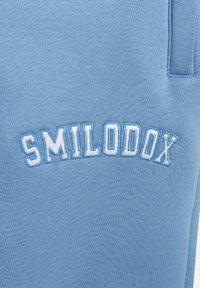 Jasnoniebieska tkanina o prążkowanej strukturze, z haftowanym słowem "SMILODOX" w białym kolorze, wygiętym u góry. Luźny styl.