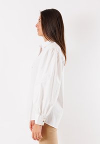 Camicia bianca di cotone con colletto button-down, maniche a sbuffo e bottoni ai polsini, esposta su uno sfondo neutro.