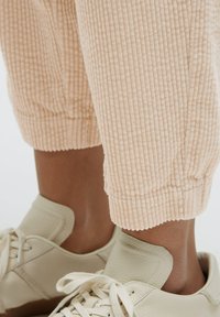 Helle beigefarbene Cord-Jogginghosen mit gerippter Textur und gerafften Bündchen, kombiniert mit beigen Low-Top-Sneakers mit Schnürdesign und runden Zehen.