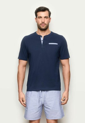 Mężczyzna ubrany w granatową krótką koszulę typu henley i białe szorty w niebieską kratę, stojący na jednolitym jasnym tle.