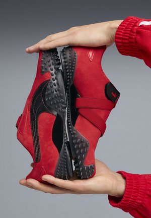 Two red and black sneakers held sole-to-sole by hands wearing red long sleeves against a gray background.