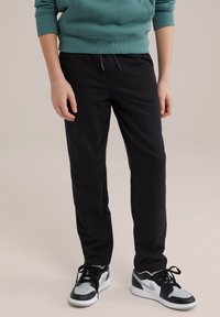 Črne trenirke z elastičnim pasom in vrvico, kombinirane z sivimi in črnimi supergami. Tkanina se zdi mehka in rahlo teksturirana.