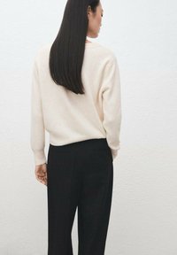 Suéter beige claro de manga larga con dobladillo acanalado, combinado con pantalones negros. El fondo es una pared blanca con textura.