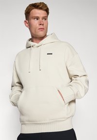 Crème-kleurige hoodie met een voorzijde kangoeroezak, ribgebreide manchetten en zoom, voorzien van een klein zwart logo op de borst. Zachte textuur.