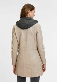 Manteau en cuir beige avec capuche noire matelassée amovible, coutures caractéristiques et taille ceinturée. Texture lisse, coupe mi-longue.