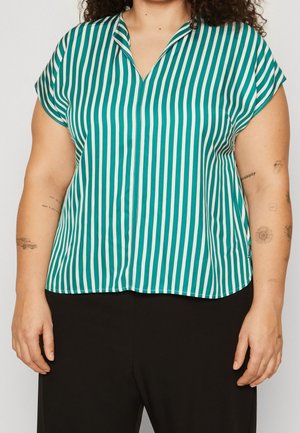 Femme portant un chemisier à manches courtes rayé verticalement vert et blanc avec un col en V et un pantalon noir, affichant des tatouages sur les deux bras.
