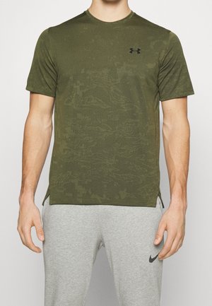 Mann trägt olivgrünes, camouflagemustertes Unter Armour Kurzarmshirt und hellgraue Nike Jogginghose, steht mit entspannten Armen.