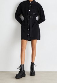 Persoon in een zwart knoopjurk met riem en borstzak, gecombineerd met zwarte stevige veterschoenen tot de enkel, staand tegen een effen achtergrond.