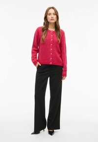 Roza pletena jopica z zapiranjem na gumbe, dolgi rokavi in rebrasto teksturo, ujemajoča se z črnimi širokimi hlačami in črnimi čevlji s koničastim nosom.