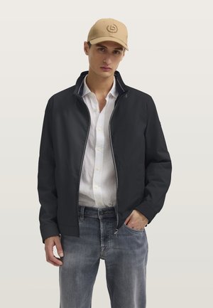 Mann trägt eine beige Kappe, schwarze Reißverschlussjacke über einem weißen Hemd mit Knöpfen und ausgebleichte blaue Jeans, steht mit einer Hand in der Tasche.