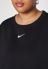 Črna dukserica s okroglim izrezom in rebrastimi manšetami, z majhnim belim Nike logotipom na sredi. Vidna je mehka tekstura tkanine.