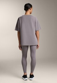 Voľné sivé tričko s krátkym rukávom v kombinácii so sivými legínami rovnakej farby. Tkanina má hladkú textúru. Čierne športové topánky dopĺňajú outfit.