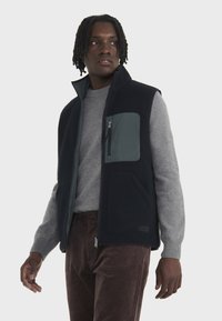 Gilet en polaire bleu marine avec une poche grise, doté d'une fermeture éclair intégrale et de poches latérales, porté par-dessus un pull gris clair et un pantalon en velours côtelé marron.