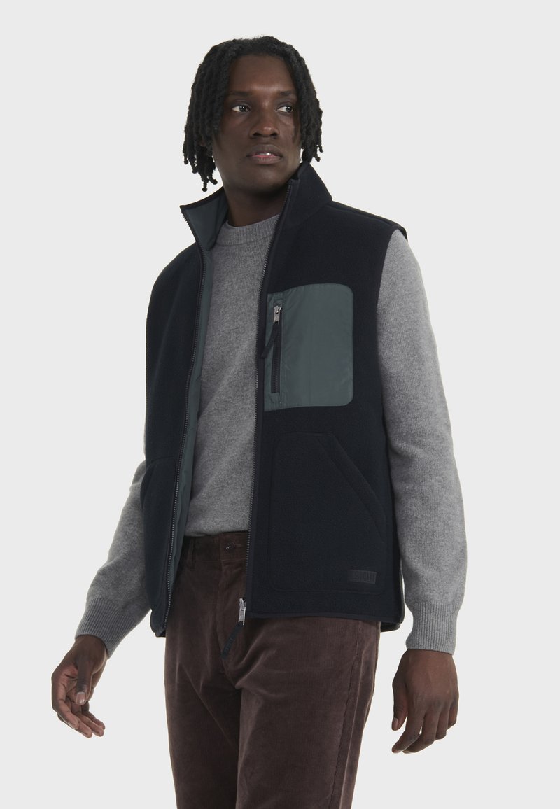 Gilet en polaire bleu marine avec une poche grise, doté d'une fermeture éclair intégrale et de poches latérales, porté par-dessus un pull gris clair et un pantalon en velours côtelé marron.