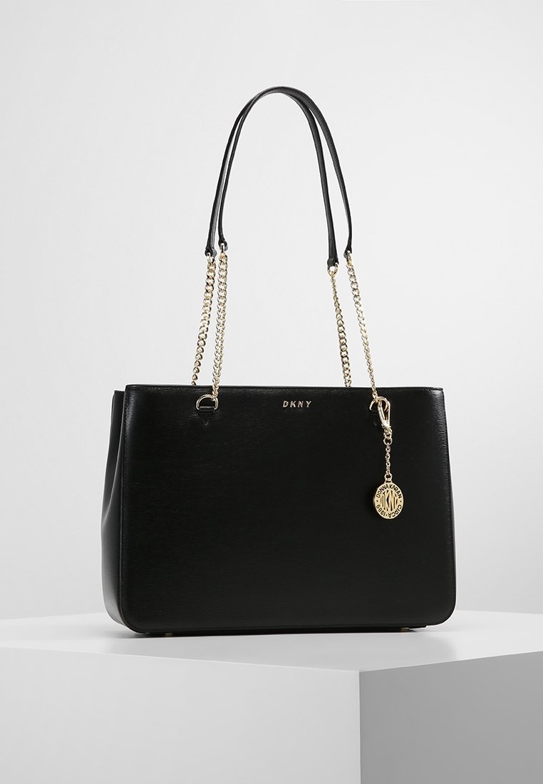 Sac cabas noir DKNY avec finition texturée, anses en chaîne dorée, détails du logo et un pendentif accroché. Forme rectangulaire avec bords lisses.
