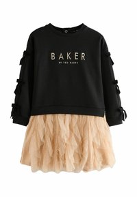 Musta huppari, jossa on kultainen "BAKER"-logo, hihoissa nauha-koristeet; kiinnitetty beige tüllihame, jossa on kerroksellinen, röyhelöinen rakenne.