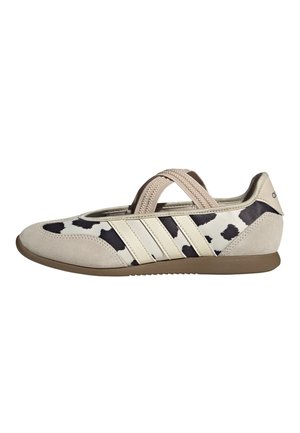 Flacher beiger Sneaker mit schwarz-weißem Kuhmuster, elastischen Kreuzriemen, drei beigen Seitenstreifen und einer braunen Gummisohle.