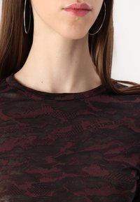 Närbild av en ung kvinnas nacke som bär en genomskinlig, bordeauxmönstrad skjorta. Stora silverörhängen kompletterar looken.