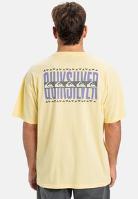 Žluté bavlněné tričko s grafickým designem na zádech, obsahujícím text "Quiksilver", trojúhelníkové vzory a symboly vln v šedé a purpurové barvě.