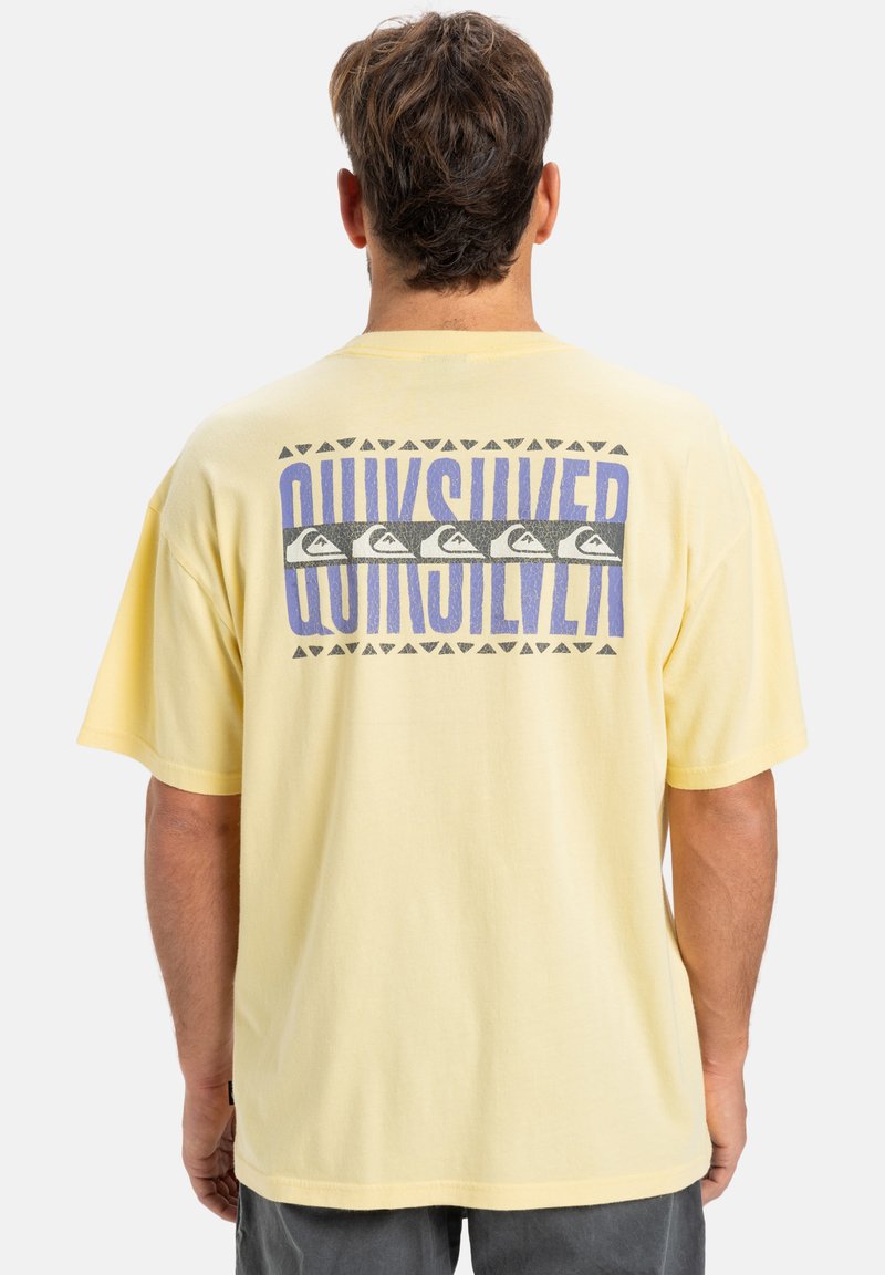 Žluté bavlněné tričko s grafickým designem na zádech, obsahujícím text "Quiksilver", trojúhelníkové vzory a symboly vln v šedé a purpurové barvě.