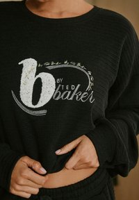 Nahaufnahme einer Person in einem schwarzen gerippten Pullover mit "b by Ted Baker" auf der Brust gestickt, die mit den Händen den Saum anpasst.