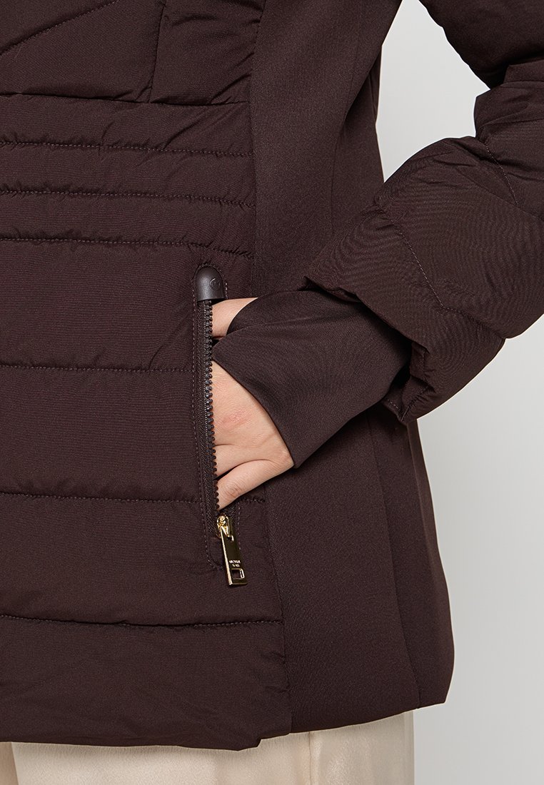 Hand in zijrits zak van een donkerbruine gewatteerde jas met een opgerolde mouw over beige broek op een lichte achtergrond.