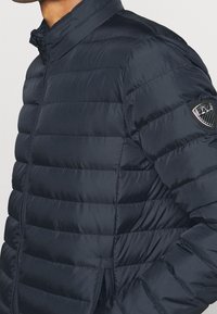 Marinblå puffjacka med horisontella sömmar, hög krage och en logotyp på övre v änstra ärmen. Slät, blänkande material.