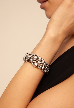 Brazalete plateado con cuentas envuelto alrededor de la muñeca, con un primer plano del brazo y una vista parcial de una persona con una manga negra.