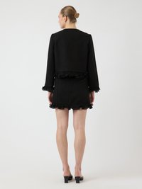 Schwarze Stoffjacke mit rüschenbesäumtem Saum und Ärmeln, kombiniert mit einem passenden Rock mit rüschenbesäumtem unteren Rand. Hohe Schuhe vervollständigen das Ensemble.