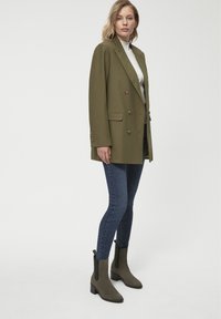 Oliwkowa dwurzędowa marynarka z dużymi guzikami, zestawiona z ciemnymi dżinsami i botkami za kostkę. Gładka tekstura, zdefiniowany krój i dopasowany fason.