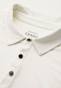 Polo shirt color crema con colletto appuntito, dotato di bottoni neri e un'etichetta con la scritta "EXIMIO" all'interno del colletto. Texture liscia.