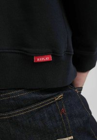 Felpa nera con orlo a costine e etichetta rossa "REPLAY". Abbinata a jeans in denim scuro con cuciture gialle e una tasca posteriore visibile.