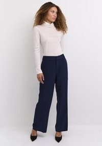 Witte, lange mouwen turtleneck van zachte stof; gecombineerd met marineblauwe wijde broek met voorplooien en een getailleerde pasvorm; zwarte hakken.