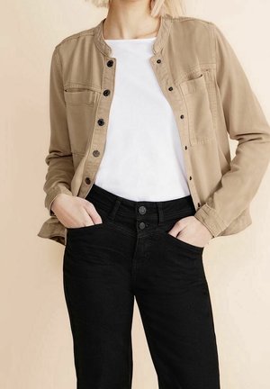 Personne aux cheveux blonds courts portant une veste beige boutonnée sur une chemise blanche et un jean noir taille haute, les mains dans les poches.