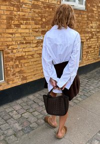 Camicia bianca a maniche lunghe con ampi polsini, gonna lavorata a maglia marrone, borsa intrecciata marrone e pantofole pelose color cammello; muro di mattoni sullo sfondo.