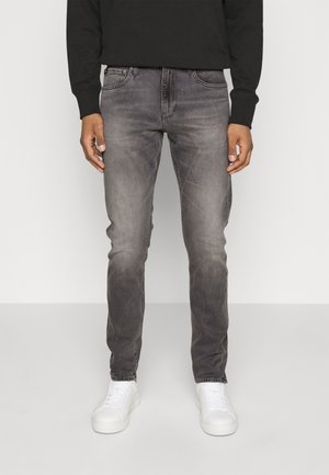 Mand iført falmede grå slim-fit jeans, sort langærmet top og hvide sneakers, stående mod en ensartet lys baggrund.