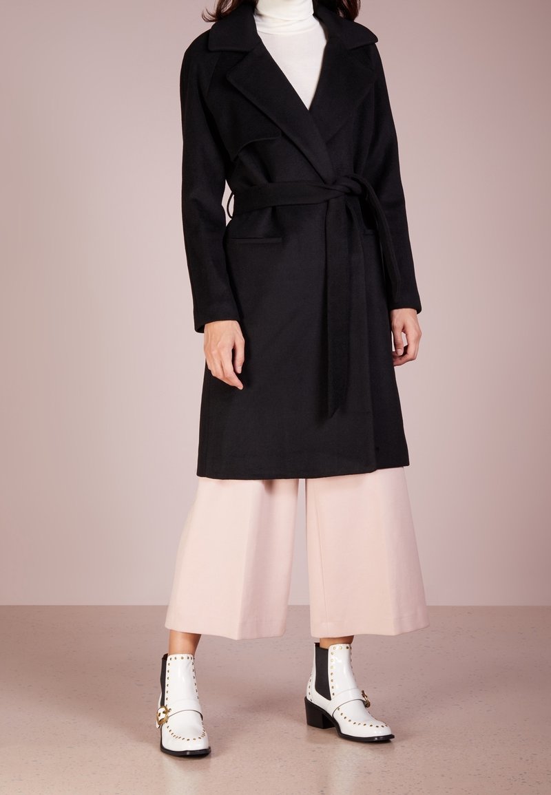Femme portant un manteau noir ceinturé sur un pantalon blanc à jambes larges et des bottines blanches avec des clous dorés et des boucles.