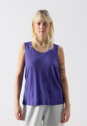 Žena s blond vlasmi, oblečená v fialovom tielku bez rukávov a svetlosivých nohaviciach, stojaca pred jednoduchým bielym pozadím.