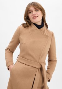 Beige wikkeljas gemaakt van zachte stof, met een hoge kraag, lange mouwen, zijzakken en een stoffen riem in de taille.