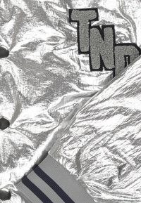 Sølv oppustelig jakke med krøllet tekstur, sorte trykknapper og "TND" broderet mærke, grå ribbede manchetter med marineblå striber.