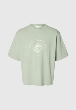 Svijetlozeleno oversized majica s bijelim kružnim grafikonom i francuskim tekstom "les pieds dans l'eau" u sredini na prsima.