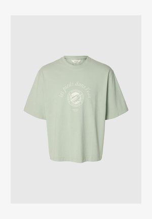 Lysegrønn oversized T-skjorte med hvit sirkulær grafikk og fransk tekst «les pieds dans l'eau» sentrert på brystet.