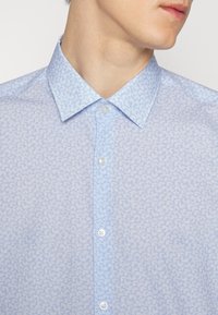 Homme portant une chemise bleu clair avec un petit motif floral blanc et un col boutonné, vu de profil.