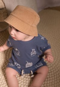 Combinaison en coton bleu avec des illustrations blanches, assortie à un chapeau seau beige. Texture douce, posée sur une surface beige texturée.