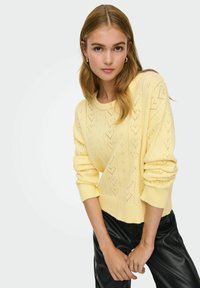 Pull jaune pâle en tricot avec un motif texturé en forme de cœur et des poignets larges côtelés, associé à un pantalon noir en simili cuir.